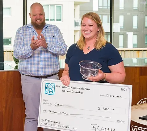 Sarah K. poses with her check for winning the 2022 Taylor Kirkpatrick Book Collecting Prize, while Taylor stands behind her and claps.